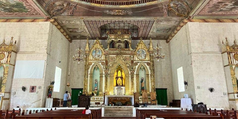 A richly decorated church interior with an ornate altar featuring gold accents and religious statues. The ceiling has intricate paintings, reminiscent of a spiritual geotrail. The pews are arranged neatly in rows. Two people are visible near the altar, one standing and one partially obscured.