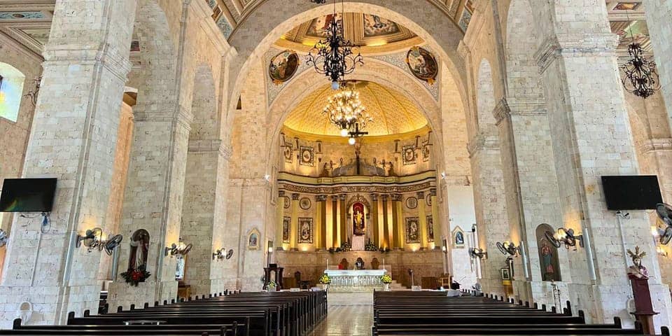 A grand cathedral interior with high arched ceilings made of stone. The altar, representing the essence of Faith, is adorned with religious statues, paintings, and a large crucifix. Pews line the central aisle leading up to the altar. Elegant chandeliers and wall sconces illuminate the space warmly.