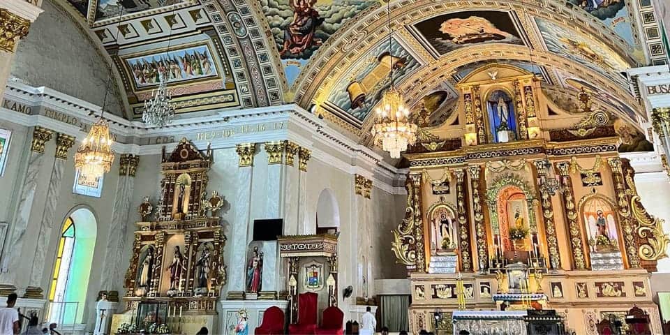 This image shows the ornate interior of a church with intricate gold and white architectural details. The ceiling and walls are adorned with religious paintings, statues, and chandeliers. The altar area, showcasing Geotrail Faith, is prominently decorated and features several religious figures.