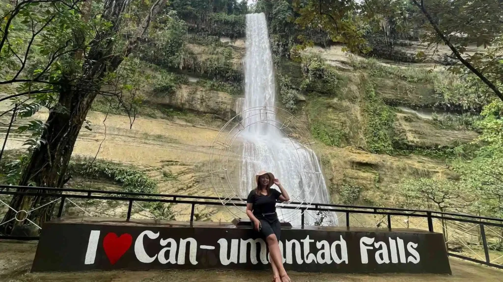 Anda-Candijay Tour - A person poses sitting on a sign that reads "I ❤️ Can-umantad Falls" with a tall, cascading waterfall in the background surrounded by lush greenery. The scene is serene and natural, emphasizing the beauty of the waterfall and its surroundings.