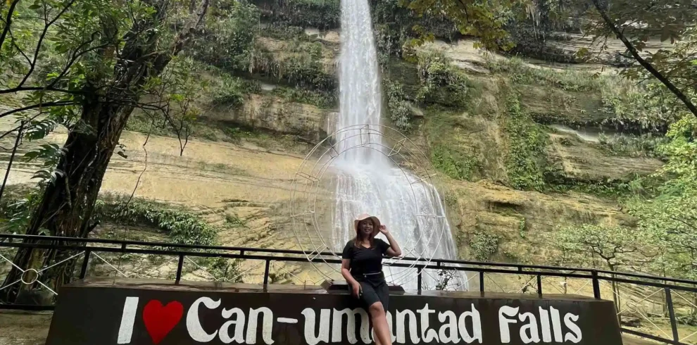 Anda-Candijay Tour - A person poses sitting on a sign that reads "I ❤️ Can-umantad Falls" with a tall, cascading waterfall in the background surrounded by lush greenery. The scene is serene and natural, emphasizing the beauty of the waterfall and its surroundings.