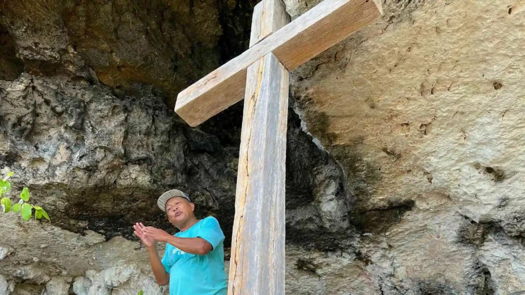 Anda-Candijay Tour - A person in a blue shirt and beige cap stands next to a large wooden cross in front of a rocky cliff. The person appears to be clapping. The cross is made of rough-hewn wood and is positioned prominently in the foreground.