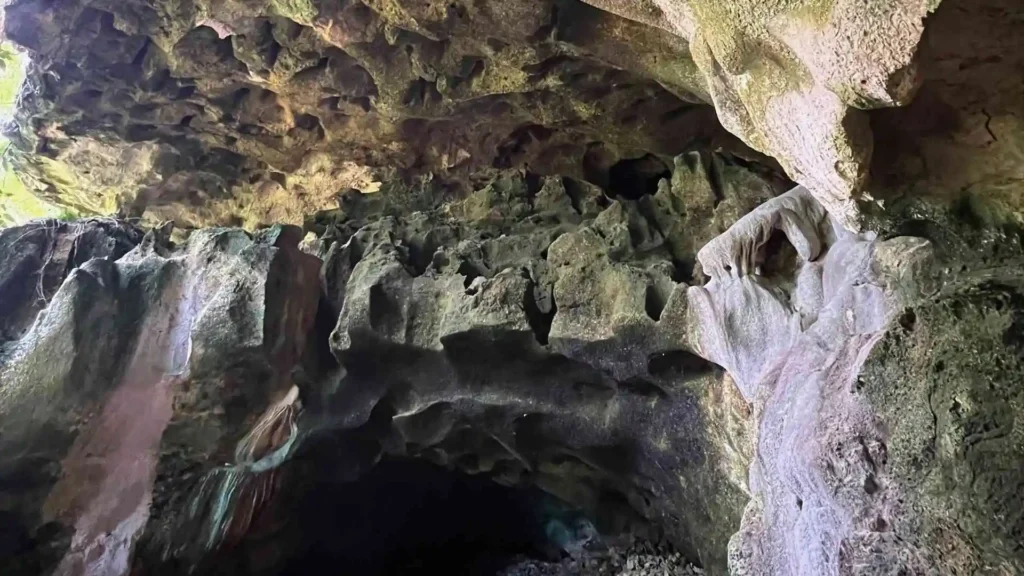 Anda-Candijay Tour - A rugged cave entrance with jagged, uneven rock formations. The light from outside highlights the textured surfaces of the rocks, revealing various shades of green and brown. The interior of the cave appears dark and mysterious.