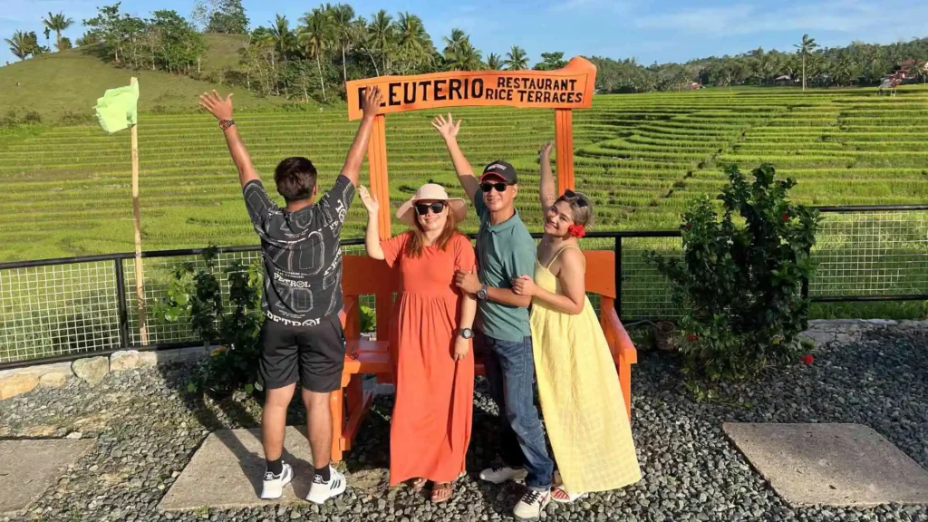 Anda-Candijay Tour - Four people are posing in front of a large wooden chair with a sign that reads "EUTERIO Restaurant Rice Terraces." They are smiling and gesturing happily. Behind them are lush green rice terraces and a clear blue sky.