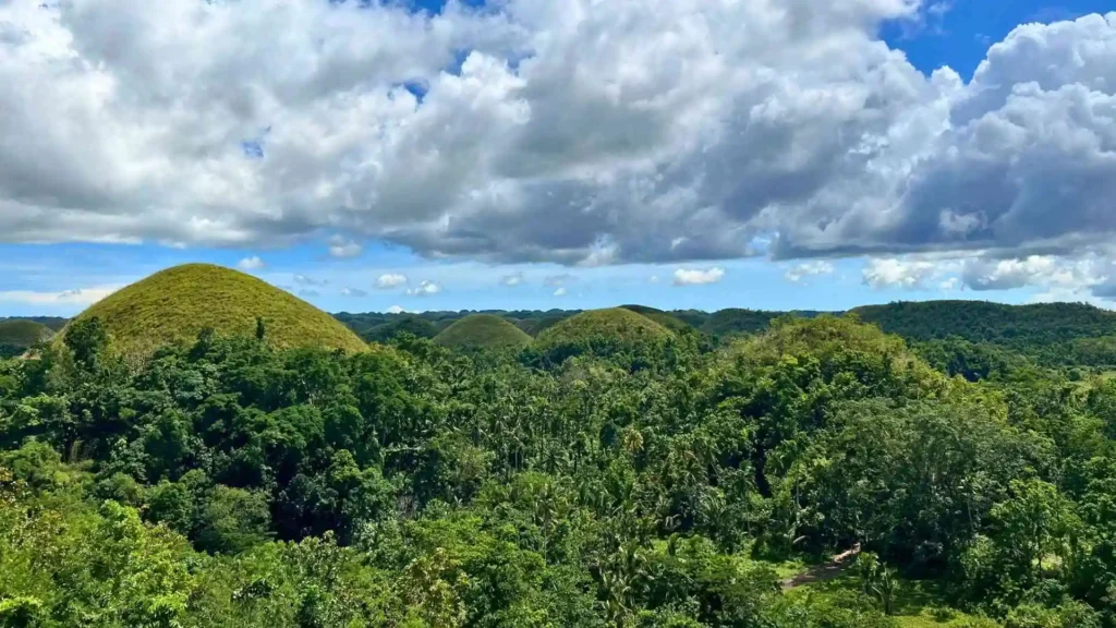 Countryside Tour - A scenic view of the Chocolate Hills, a series of lush green hills under a partly cloudy sky in Bohol, Philippines. Dense foliage covers the surrounding area while the hills themselves have a unique, conical shape.