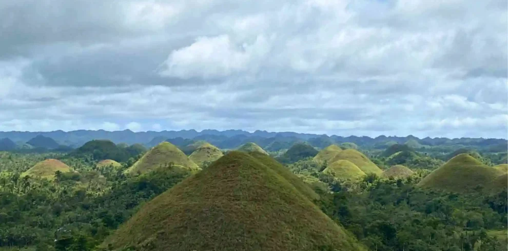 Countryside Tour - A scenic view of the Chocolate Hills, characterized by numerous green, grassy hills under a partly cloudy sky. The mounds rise uniformly from a dense, lush forest, creating a captivating, undulating landscape stretching to the horizon.