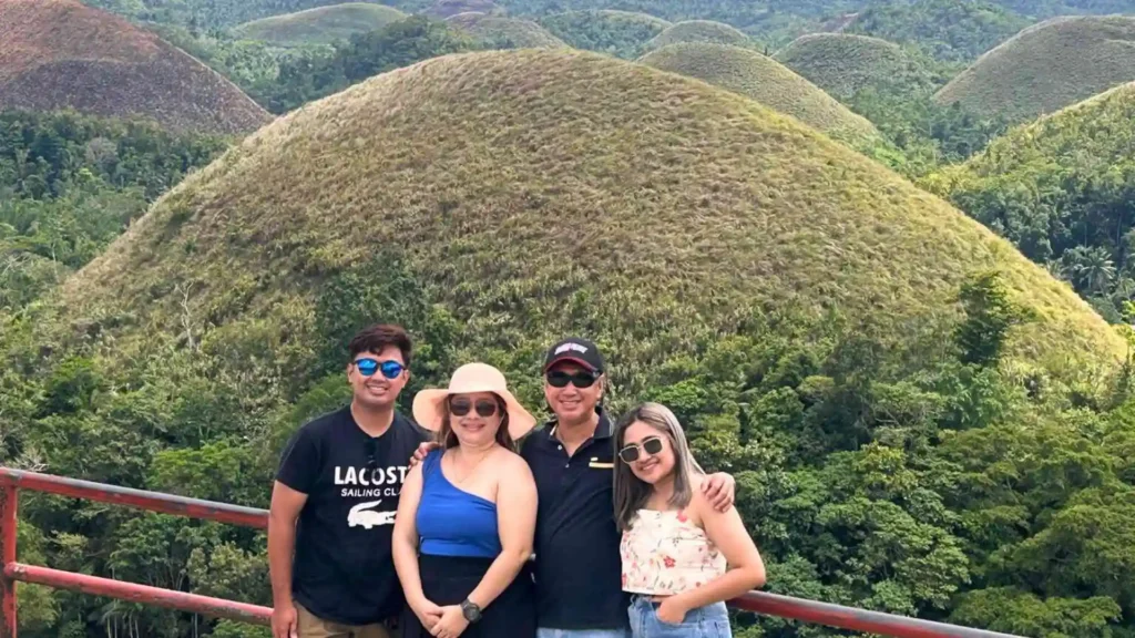 Countryside Tour - A group of four people, two men, and two women, posing together in front of the Chocolate Hills in Bohol, Philippines. They are standing on a wooden platform with lush green hills in the background, all smiling and wearing casual outdoor clothing.