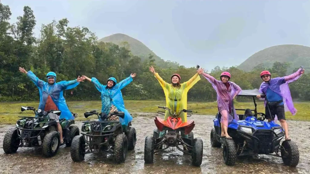 Countryside Tour - A group of five people dressed in colorful raincoats pose joyfully with their arms raised while sitting on all-terrain vehicles (ATVs). They're on a muddy trail with lush green trees and a mountain visible in the background, under overcast skies.