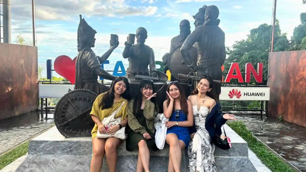 Countryside Tour - Four women are sitting on a stone bench, smiling for a photo with large statues of historical figures in the background. The statues depict individuals raising mugs in a toast. The women are wearing casual summer outfits. The background includes colorful letters and signage.