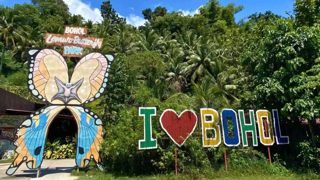 Countryside Tour - The image shows the entrance to the Bohol Butterfly Park. The entrance is adorned with a large butterfly structure. Next to it is a colorful sign reading "I ❤️ Bohol" against a backdrop of lush green palm trees and foliage.