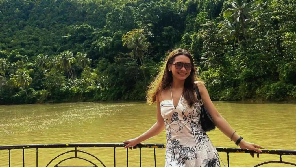 Countryside Tour - A woman in a sleeveless dress and sunglasses stands smiling next to a railing with a river and lush green forest in the background. She is holding onto the railing with one hand, and the scene is bright and sunny.