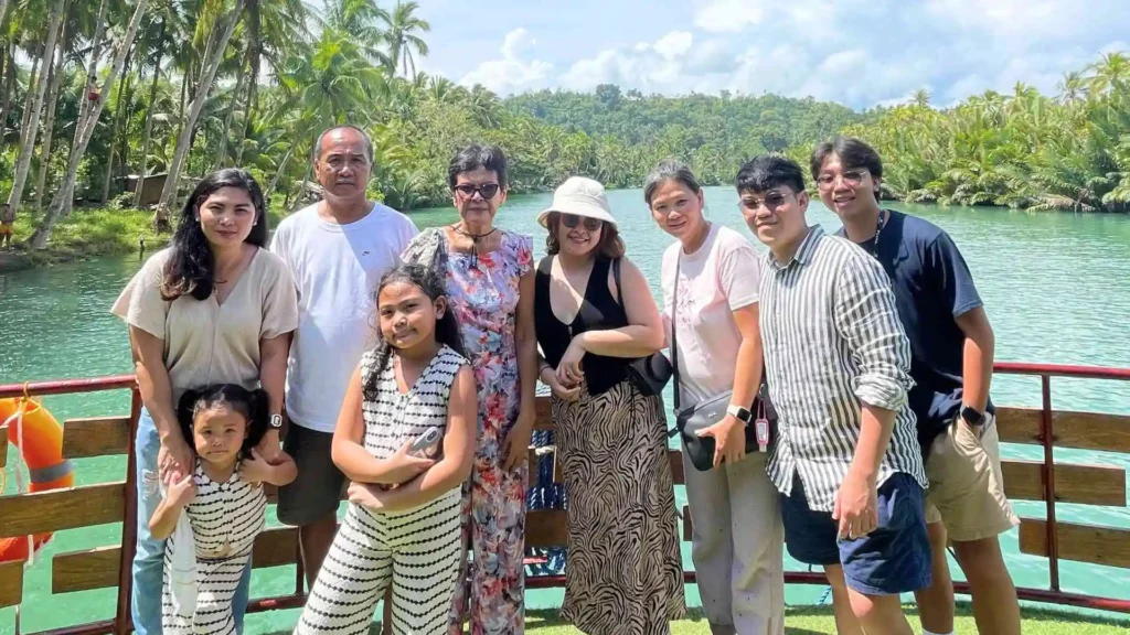 Countryside Tour - A group of ten people, including adults, teenagers, and children, stands together outdoors on a sunny day. They are posing in front of a lush, tropical background with palm trees and a river. Some are wearing sunglasses and summer attire.