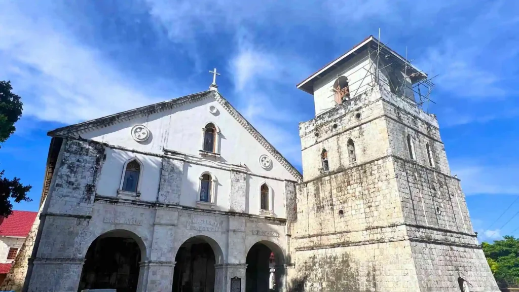 Faith-Based Geotrail Tour - A historical stone church with a white façade and arched entrances stands prominently under a bright blue sky. Adjacent to it is a tall, square bell tower with visible scaffolding. The architecture showcases a mix of colonial and baroque styles.