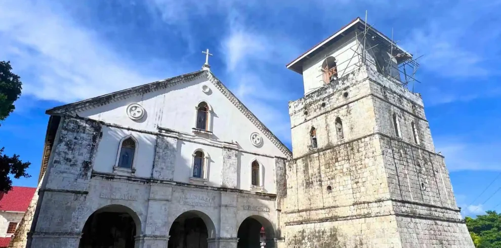 Faith-Based Geotrail Tour - A historical stone church with a white façade and arched entrances stands prominently under a bright blue sky. Adjacent to it is a tall, square bell tower with visible scaffolding. The architecture showcases a mix of colonial and baroque styles.