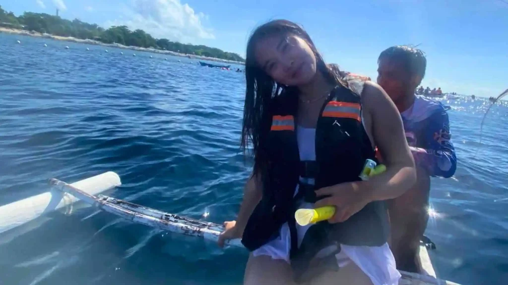 Island Hopping Tour - A young woman wearing a life jacket smiles while sitting in a small boat on a sunny day. She appears to be holding snorkeling gear. The blue water around her is calm, with the shoreline visible in the background. Another person is seated behind her in the boat.