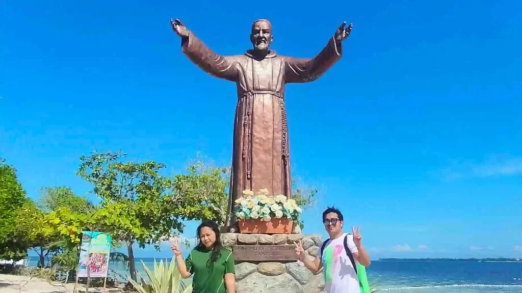 Island Hopping Tour - Two individuals stand in front of a large statue of a robed figure with arms outstretched. The statue is situated outdoors with a clear blue sky and a glimpse of the ocean in the background. Both individuals are smiling and making peace signs with their hands.