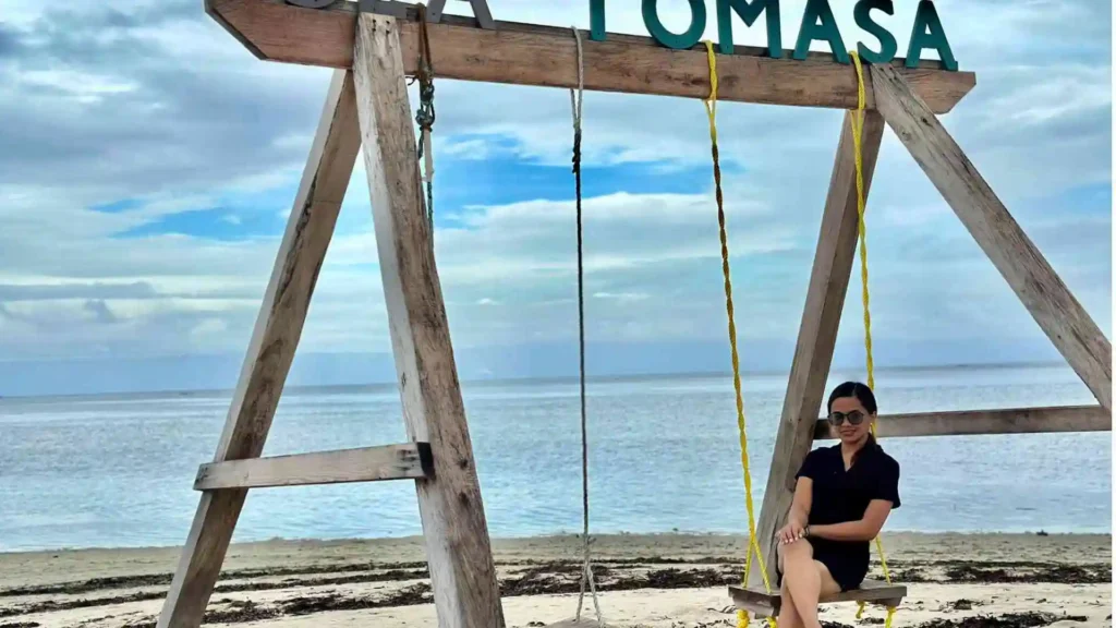 Island Hopping Tour - A person in a black outfit with sunglasses is sitting on a yellow swing attached to a wooden structure labeled "VILLA TOMASA." The swing is set on a sandy beach with a calm sea and a blue sky with some clouds in the background.