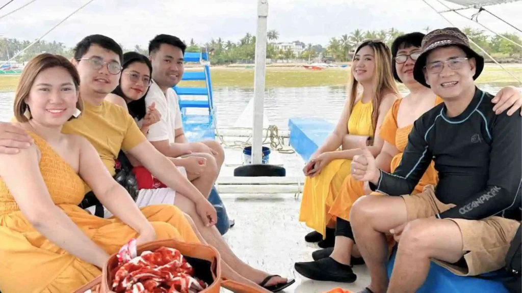 Island Hopping Tour - A group of seven people are sitting on a boat, smiling at the camera. The background features a calm body of water and a distant shoreline with trees and structures. Several individuals are wearing yellow outfits. One person on the right is giving a thumbs-up.