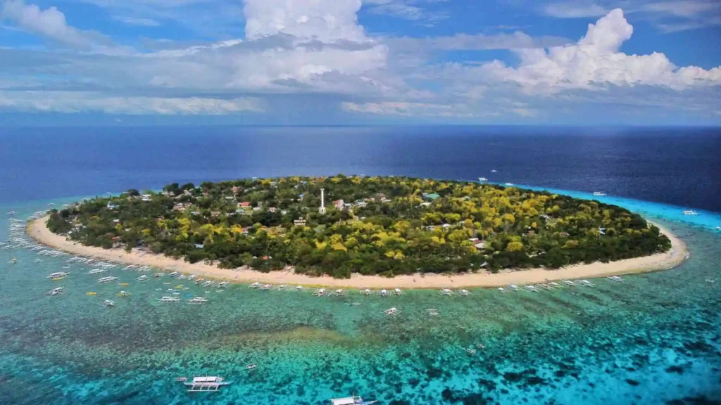 Island Hopping Tour - Aerial view of a lush, circular island surrounded by turquoise waters and coral reefs. Sandy beaches border the island, with various boats and small structures visible along the shoreline. The sky is partly cloudy, adding depth to the vibrant seascape.