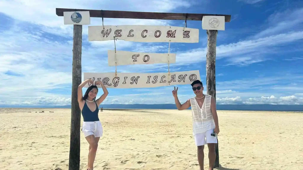 Island Hopping Tour - Two people pose under a wooden sign that reads "Welcome to Virgin Island" on a sandy beach, with a clear blue sky and ocean in the background. The person on the left waves while the person on the right shows a peace sign.