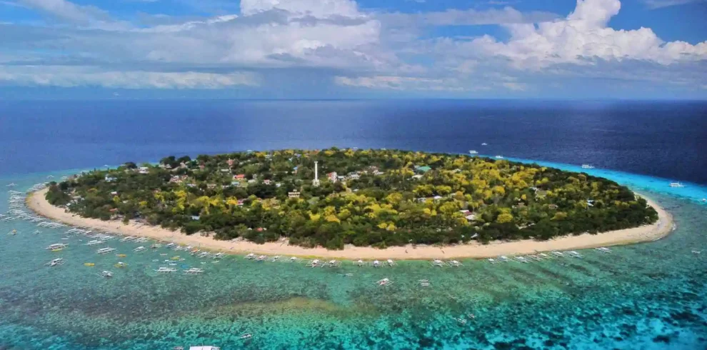 Island Hopping Tour - Aerial view of a lush, circular island surrounded by turquoise waters and coral reefs. Sandy beaches border the island, with various boats and small structures visible along the shoreline. The sky is partly cloudy, adding depth to the vibrant seascape.