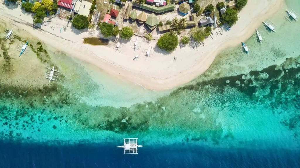 Pamilacan Island Hopping Tour -Aerial view of a coastal area featuring a gradient from sand to ocean. The shore is lined with buildings, trees, and small boats. The water changes from clear turquoise near the beach to a deep blue further out. A distinctive white boat is anchored in the blue water.