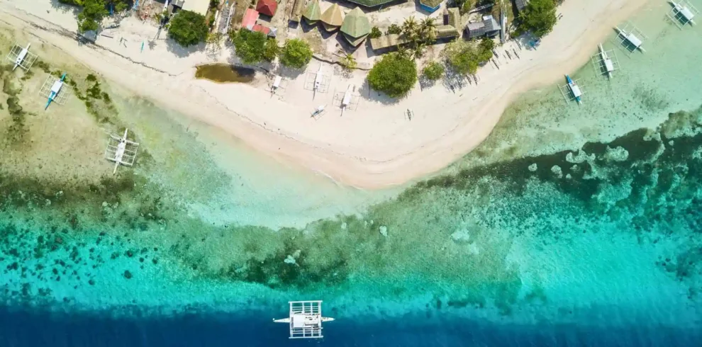 Pamilacan Island Hopping Tour -Aerial view of a coastal area featuring a gradient from sand to ocean. The shore is lined with buildings, trees, and small boats. The water changes from clear turquoise near the beach to a deep blue further out. A distinctive white boat is anchored in the blue water.