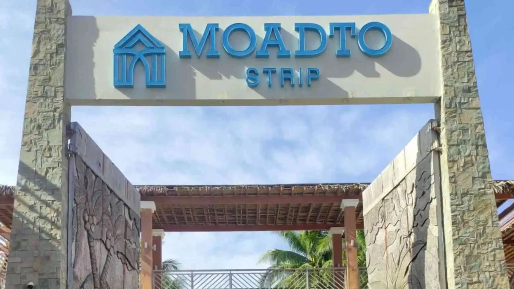 Bohol-Panglao Tour - A stone archway with the text "MOADTO STRIP" displayed prominently overhead. The path underneath leads to an outdoor area with palm trees and lattice roofing visible in the background. The sky is clear and bright.