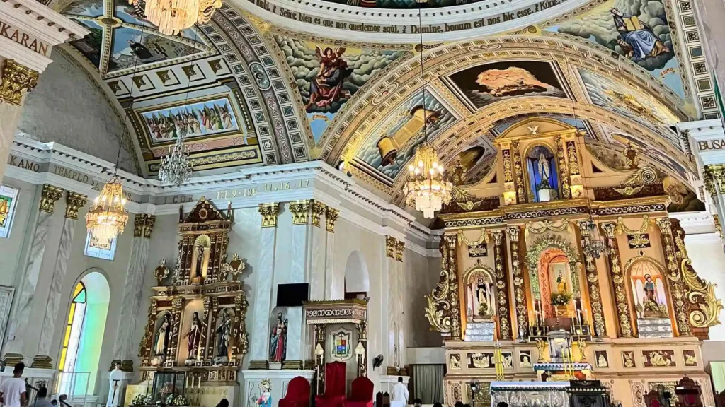 Bohol-Panglao Tour - A richly decorated church interior featuring ornate gold details, intricate wall and ceiling murals, and chandeliers. The altar area has large religious statues and elaborate designs, with pews and red chairs in the foreground. Stained glass windows are visible.