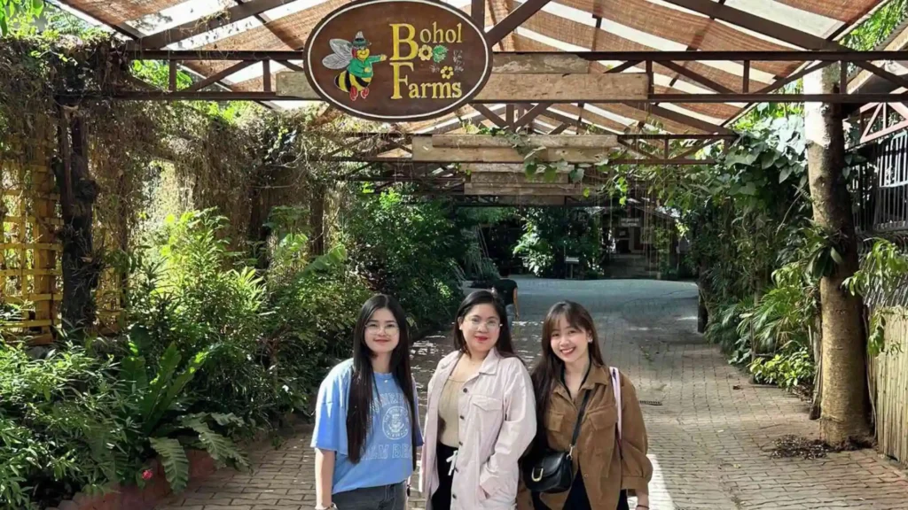 Bohol-Panglao Tour - Three people stand smiling under a wooden pergola with a sign that reads "Bohol Bee Farm". They are surrounded by lush green foliage and plants. The scene suggests they are visiting the farm as tourists.