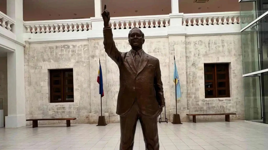 Bohol-Panglao Tour - A bronze statue of a man wearing a suit is standing with one arm raised pointing upwards. The statue is placed in an open, white-tiled hall. Behind it, there are two benches and several flags displayed against a stone wall with windows.