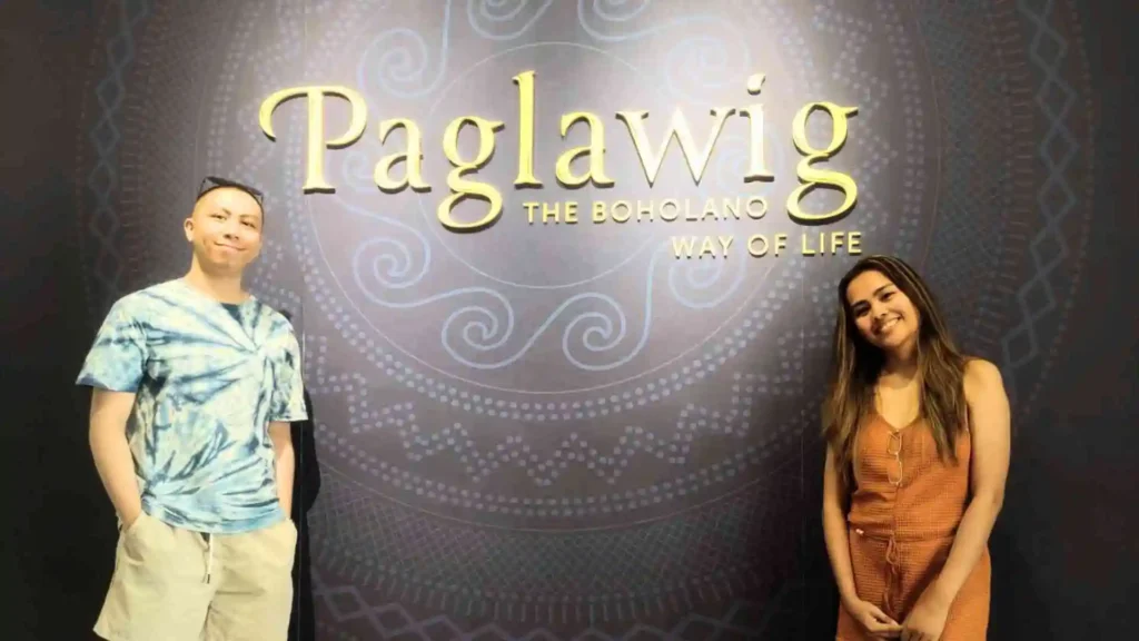 Bohol-Panglao Tour - Two people are standing and smiling in front of a decorative background with the words "Paglawig: The Boholano Way of Life" displayed prominently. The person on the left is wearing a blue tie-dye shirt and light-colored shorts, while the person on the right is in a rust-colored dress.