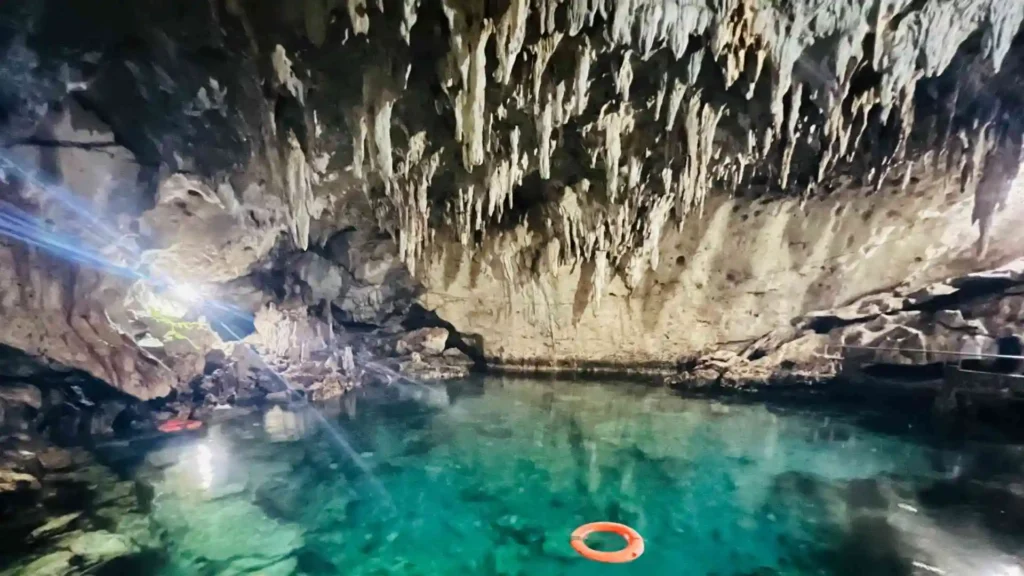 Bohol-Panglao Tour - A stunning underground cave with clear, turquoise water and dramatic stalactites hanging from the ceiling. A red life ring floats on the water, and a spotlight illuminates a rocky area to the left. The serene environment suggests a natural, secluded beauty spot.