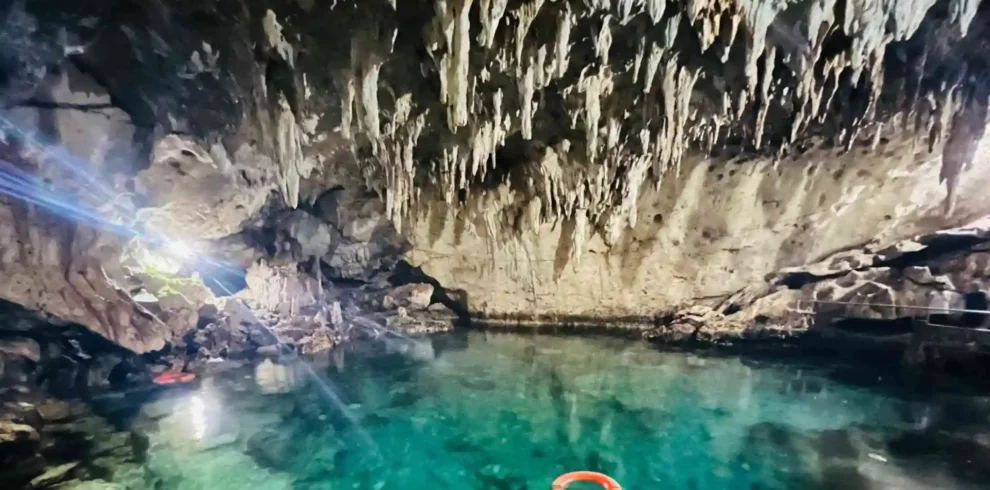 Bohol-Panglao Tour - A stunning underground cave with clear, turquoise water and dramatic stalactites hanging from the ceiling. A red life ring floats on the water, and a spotlight illuminates a rocky area to the left. The serene environment suggests a natural, secluded beauty spot.