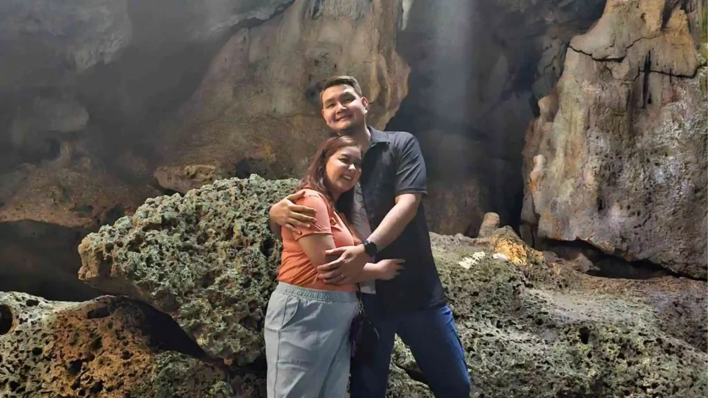 Bohol-Panglao Tour - A couple stands in a cave, embracing and smiling. They are surrounded by large rock formations with light filtering in from above, creating a serene and intimate atmosphere. The woman wears a coral top and light blue pants, while the man wears a dark shirt and jeans.