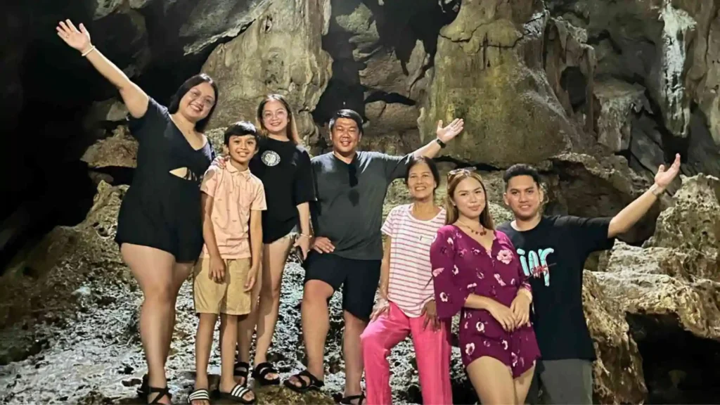 Bohol-Panglao Tour - A group of eight people stand together smiling and posing with open arms inside a cave. They are dressed casually and appear to be enjoying their time exploring the cave. The rock formations behind them are visible and the lighting is dim but adequate.