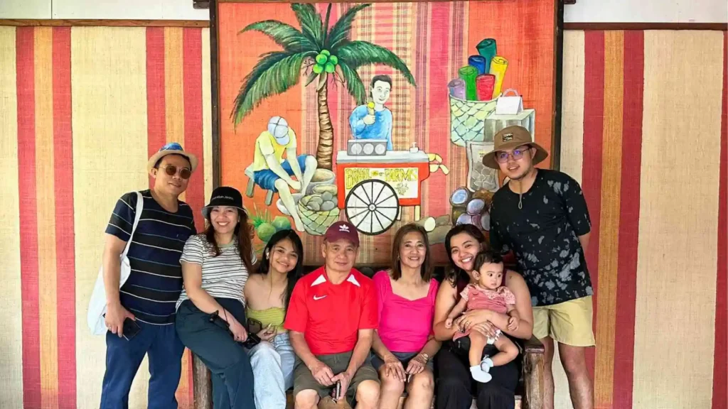 Bohol-Panglao Tour - A group of eight people, men and women, are posing together in front of a colorful mural depicting a tropical scene. One person is holding a baby. The group is dressed casually and appears to be enjoying themselves.