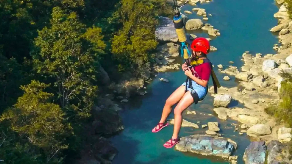 Danao Extreme Tour -Person wearing a red helmet and harness zip-lines over a rocky, turquoise river. The individual is dressed in a maroon shirt, denim shorts, and pink sandals, and is surrounded by lush green trees.