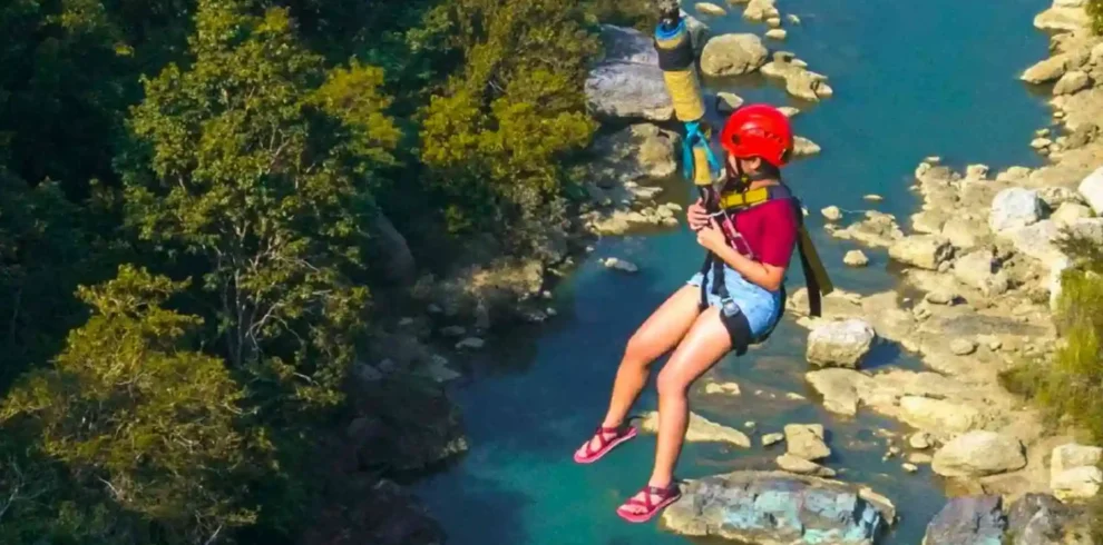 Danao Extreme Tour -Person wearing a red helmet and harness zip-lines over a rocky, turquoise river. The individual is dressed in a maroon shirt, denim shorts, and pink sandals, and is surrounded by lush green trees.