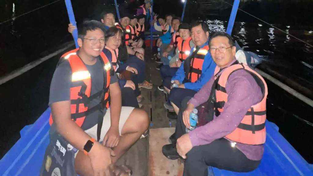 Firefly Watching Tour - A group of people wearing life jackets sit closely together on a boat at night. They appear to be on a guided tour, enjoying the ride. Some are smiling at the camera, while others look around or talk to each other. The water around them is dark, and some lights are visible.