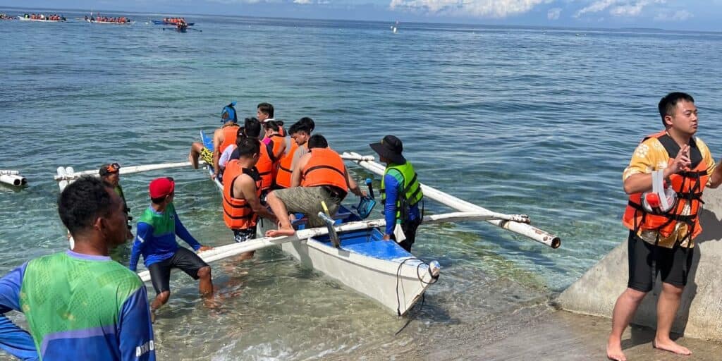 A group of people wearing life jackets is boarding a small boat at the edge of the sea. The water is clear and calm, with another boat visible in the background. The sky is clear with scattered clouds. Some people are standing on a rocky shoreline helping others board.