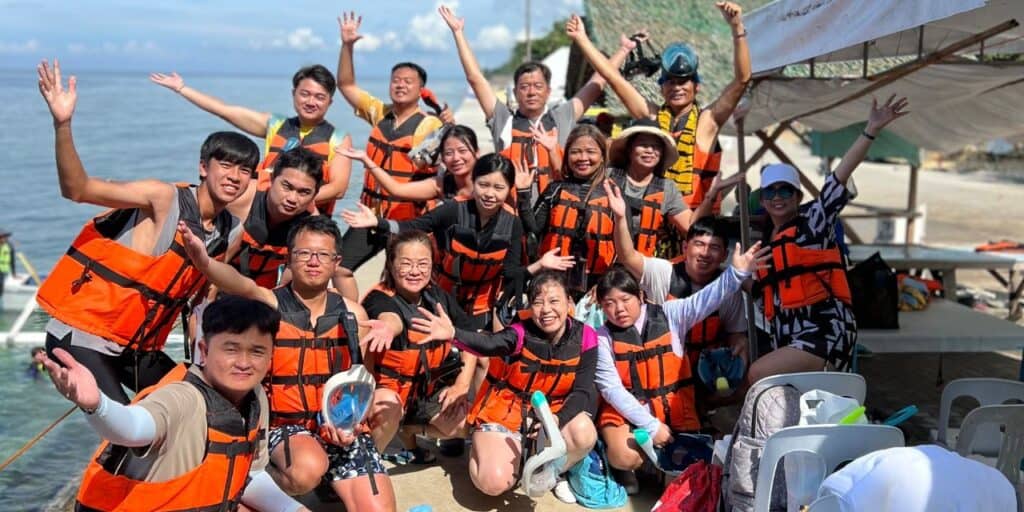 A group of people wearing orange life vests gather and cheer in front of a scenic seaside backdrop. Several members stand while others sit, all smiling and raising their hands joyfully. The scene appears festive and full of camaraderie.