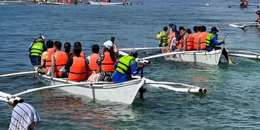 A group of people wearing orange life jackets are sitting in a white outrigger canoe on a clear blue body of water. Another group of people in green and orange life jackets are in a second canoe nearby. More people and canoes are visible in the background.